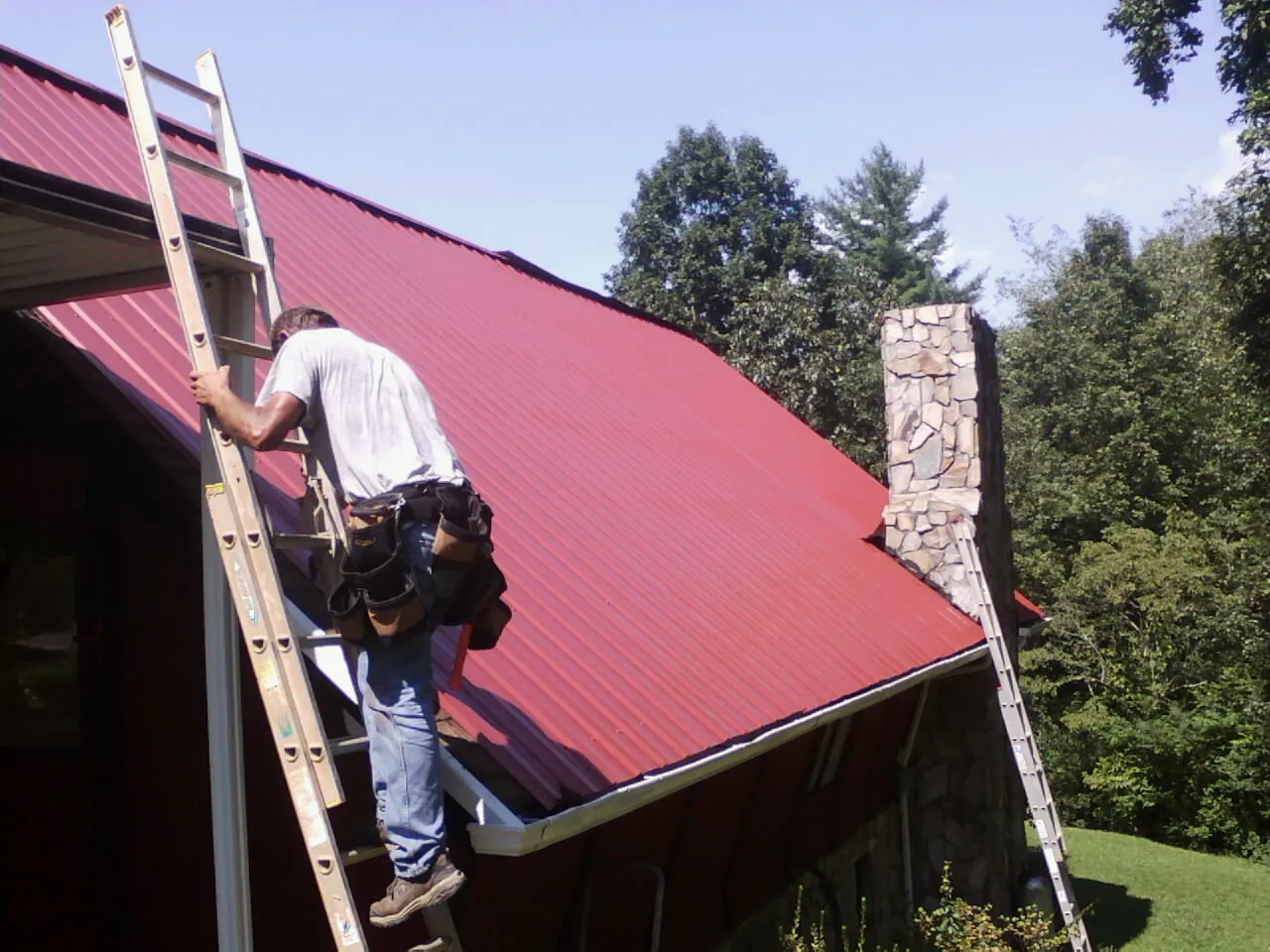 Roofer on a ladder beside a red metal roof and stone chimney — Soffit & Fascia Repair by Roofing Brothers in Black Creek